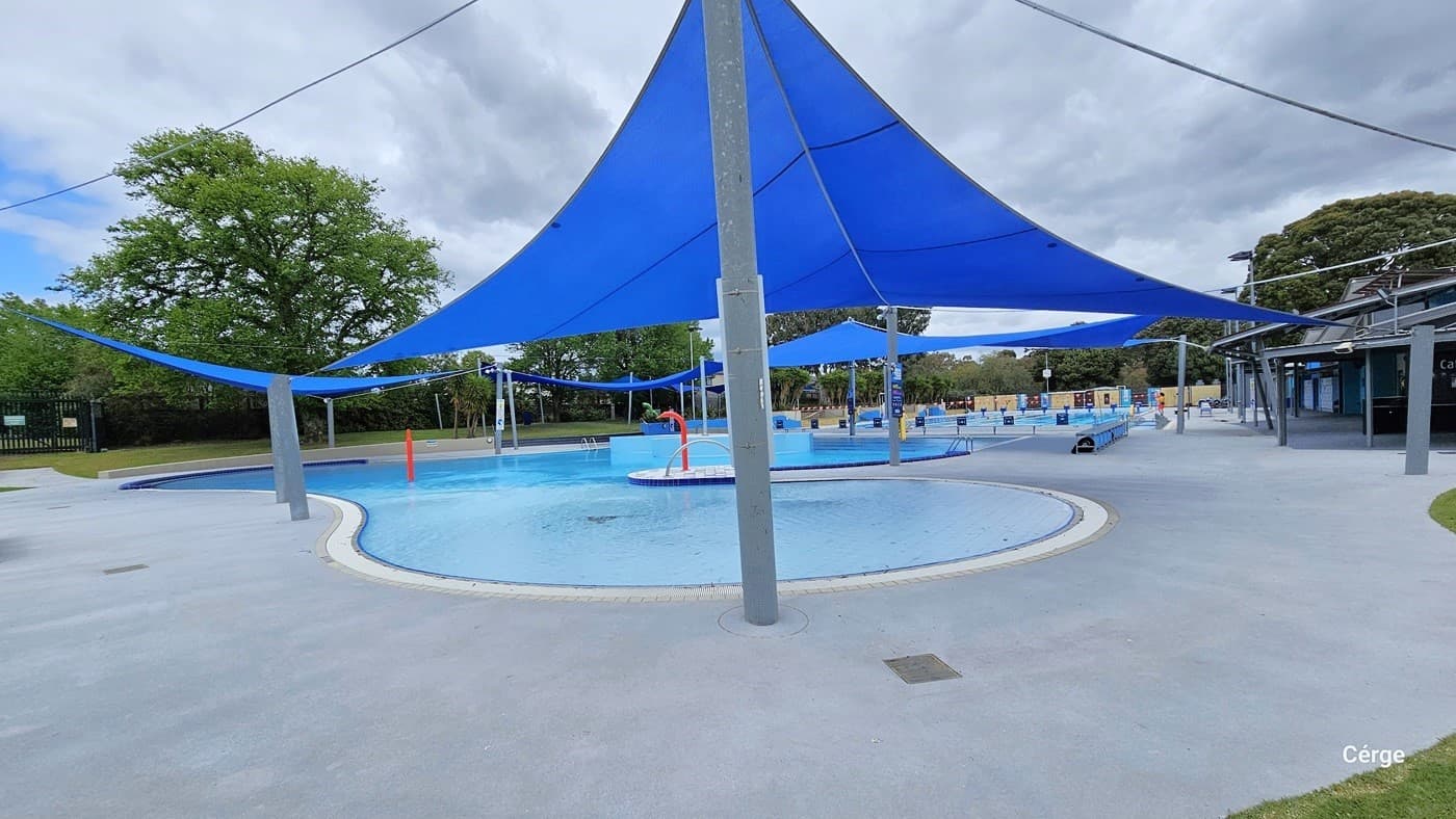 Circular shallow pool with 0.7m depth equipped with water splash features for younger swimmers. It has a white tile pole coping and a wide grey pool deck. The pools have large blue sun shades that provide shade during the day. Red metal posts with a curved end for water splash are placed in the middle and on the pool coping. The black perimeter fence, the 8-lane, 50-meter pool, and the café can be seen from this position.