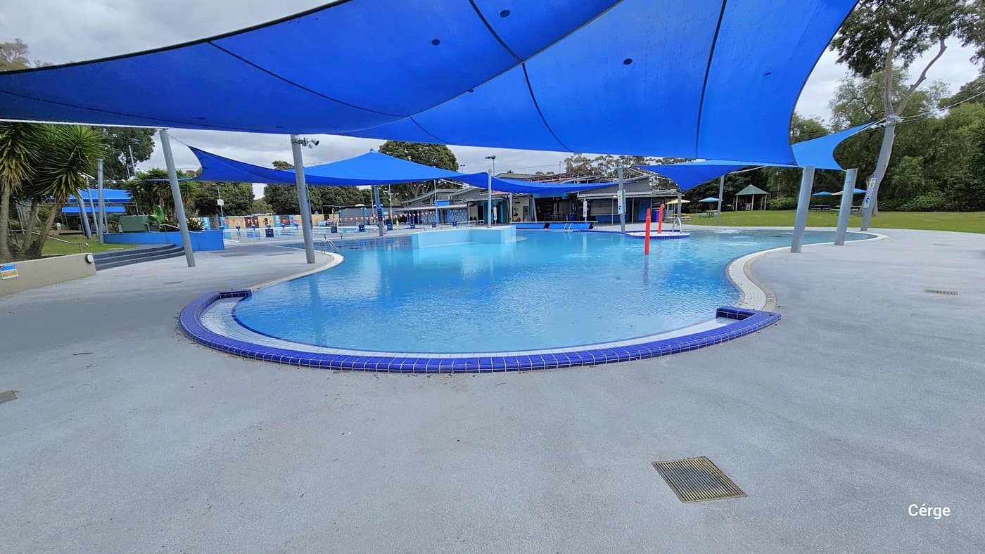 A closer look at the shallow pool with 0.7 meter depth for children. This side of the pool has a blue pool tile coping on top of the white pool tile coping. Red metal posts with a curved end for water splash are placed in the middle and the poolside. Further to the right is a grey stair with metal handrails, while beyond the café is a BBQ station with picnic tables and umbrella shades of varying colors.
