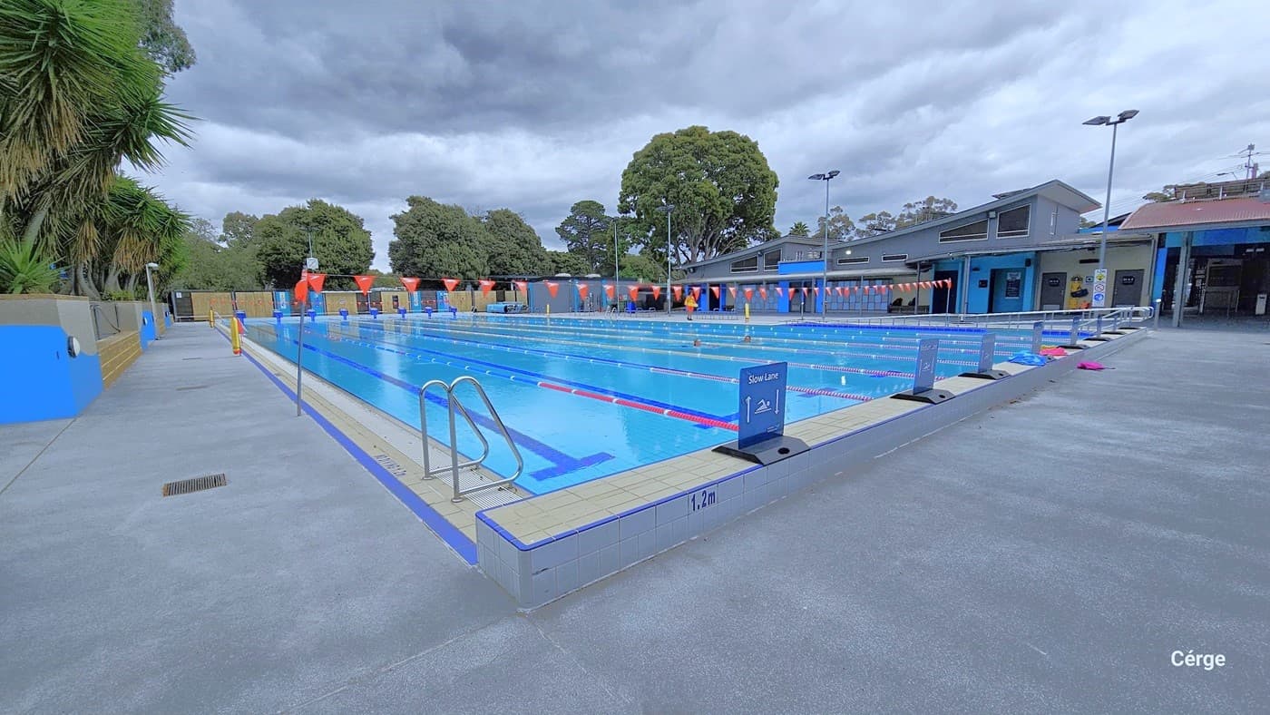 View from the 1.2m depth of the eight-lane, 50m pool. There is a pool ladder on the right white tile pool coping. Two yellow floating devices are wrapped around the grey metal posts on the right pool deck. On one end of the pool are blue signs with black flat support that has a white swimming icon and white arrows on each side for slow and fast swimming lanes. Each lane is separated by red, blue, and white floating rope lanes.