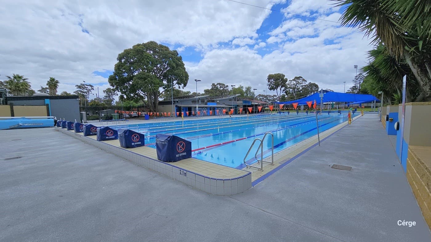View of the eight-lane, 50m pool on the 2.1-meter end. On the top of the elevated white tile coping are diving blocks covered with a blue waterproof cloth with a NO Diving sign. There is a pool ladder on this side marked by two inverted U-stain steel handles. On the right side is a concrete brick wall with gold, grey, and blue color schemes. Each lane is separated by red, blue, and white floating rope lanes. Yellow floating devices are wrapped around grey metal posts on the right pool deck.