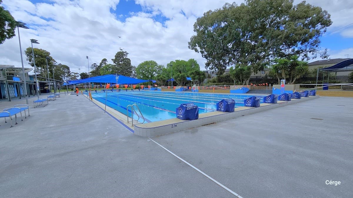 Another angle of the eight-lane, 50m pool with a 2.1-meter depth. On the top of the elevated white tile coping are diving blocks covered with a blue waterproof cloth with a NO Diving sign. There is a pool ladder on this side marked by two inverted U-stain steel handles. There are several seats with blue seat covers supported by curved stainless steel frames on the left grey pool deck. Tall lamp posts are placed slightly behind each seating.