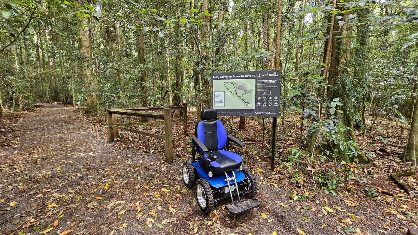 A forest path with a dirt trail covered in fallen leaves on the left. On the right, there's a blue and black mobility scooter parked in front of a sign that reads "Mary Cairncross Scenic Reserve rainforest walks." The sign includes a map and various symbols. A wooden fence is nearby, and tall trees with dense green foliage surround the area.