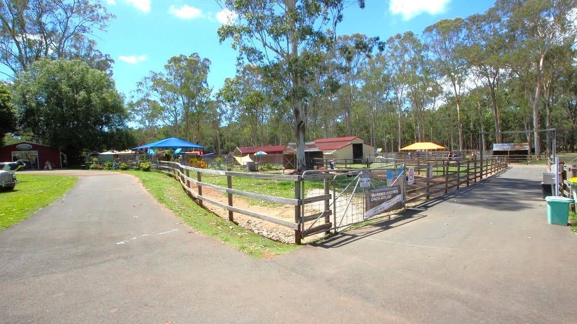 Down the path from the ticket office is the toddler playground which is in a fenced area on the right that has blue roofing. On the other left, is a manikin a cow. The gate leading to the toddler playing ground has the signage, “BARRIER ROOFING 0408 40822”. The wooden fence also houses the big barn and the party barn which both have red roofing and white walls. Right across the entrance to the toddler playing area is a green rubbish bin.