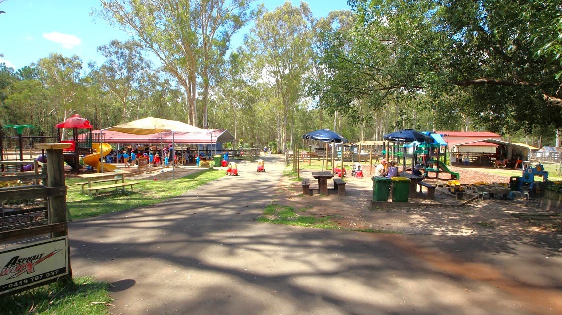 At the toddler’s play area are kids' play areas and sitting areas. The sitting areas are made of wood and are covered by blue umbrellas and some are covered under the shade of the trees. At the entrance to the play area is a sign with ‘Asphalt Worx’. The path leading to the play area is cemented with two slides on either side. There is a cream-covered pergola under which the children get to play. There is a pergola at the end of the play area that is also a rest area.