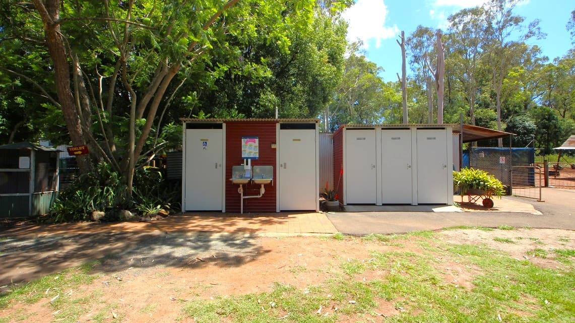There are five bathrooms at White Ridge Farm. The bathroom doors are painted white and are separated into three bathrooms on one side and two bathrooms on the other side. In between the two sets of bathrooms is a plant with red leaves in a white pot. There are two hand wash basins placed between the two bathrooms with green plants surrounding. On the left side of the bathrooms is a plant with yellow leaves at the edge.