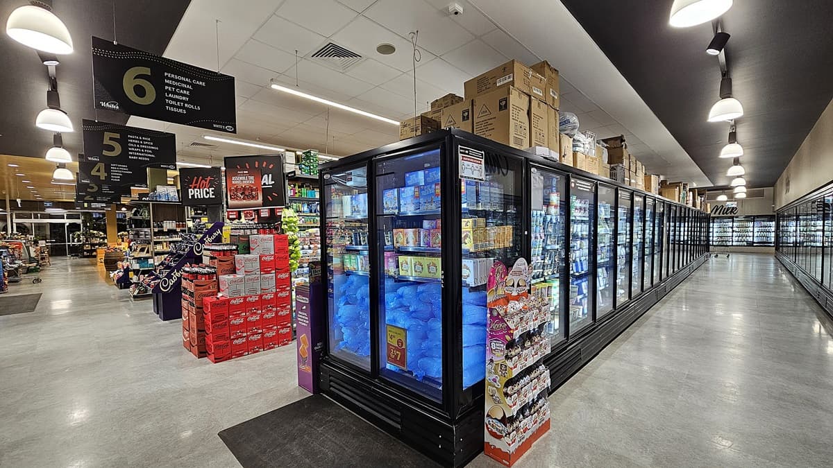 On the left, there are shelves stocked with various products, including a prominent display of red boxes and a sign indicating aisle 6. In the middle, there is a long row of refrigerated units filled with frozen goods, with some promotional items attached to the ends. On the right, more refrigerated units extend into the distance. The floor is polished concrete, and the ceiling features recessed lighting and ventilation panels.