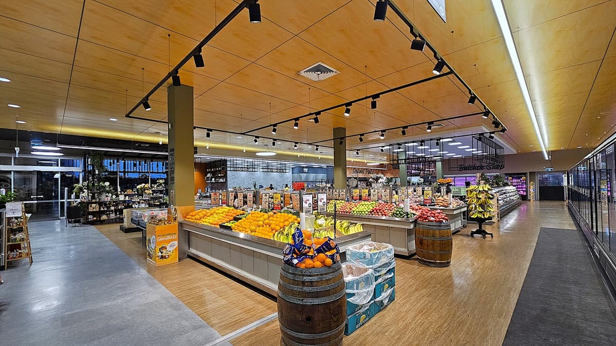 On the left, there are glass doors leading outside and a section with various plants and flowers. In the middle, there are large displays of fresh fruits and vegetables, including oranges, apples, and bananas, arranged on wooden barrels and tables. On the right, there are refrigerated units with glass doors containing dairy products. The floor is a combination of polished concrete and wood, and the ceiling is wooden with recessed lighting and ventilation panels.