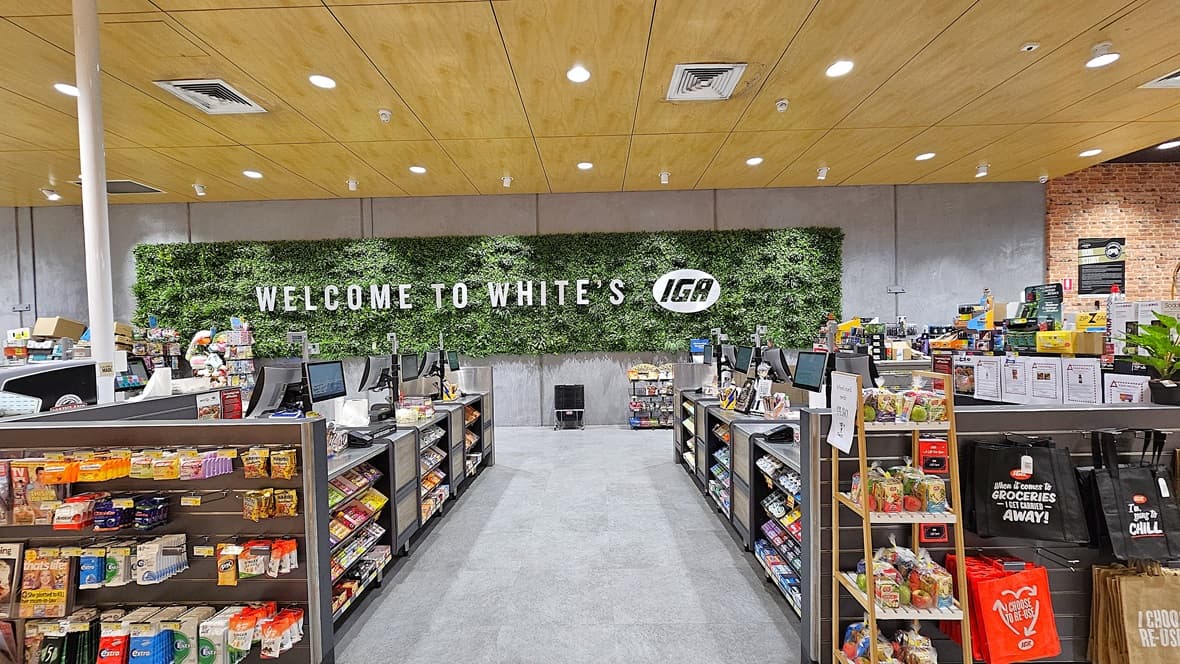 On the left, there are shelves stocked with various snacks and a display of colourful packaged goods. In the middle, there is a wide aisle leading to a wall with a large sign reading "Welcome to White's IGA" against a backdrop of green foliage. On the right, there are checkout counters with tills and more shelves filled with snacks and drinks. The floor is carpeted, and the ceiling is wooden with recessed lighting and ventilation panels.