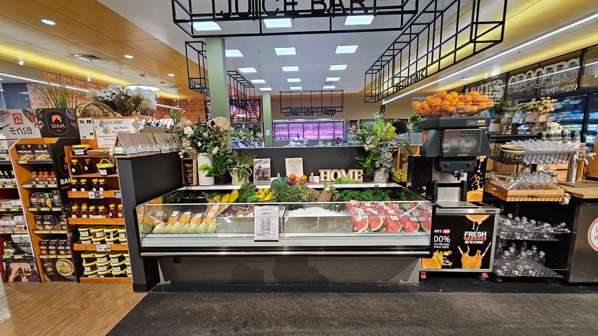 On the left, there are shelves filled with various bottles and a display of packaged goods. In the middle, there is a glass counter showcasing fresh produce, including melons and pineapples, with a "HOME" sign on top. In the background, there is a deli section with a sign reading "Butcher's Corner." On the right, there are shelves with glassware, a coffee machine, and a display of oranges. The floor is a mix of wood and carpet, and the ceiling has recessed lighting and black metal frames.