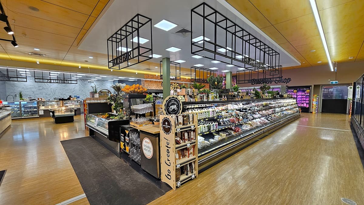 On the left, there are shelves filled with various bottles and a display of packaged goods. In the middle, there is a glass counter showcasing fresh produce, including melons and pineapples, with a "HOME" sign on top. In the background, there is a deli section with a sign reading "Butcher's Corner." On the right, there are shelves with glassware, a coffee machine, and a display of oranges. The floor is a mix of wood and carpet, and the ceiling has recessed lighting and black metal frames.