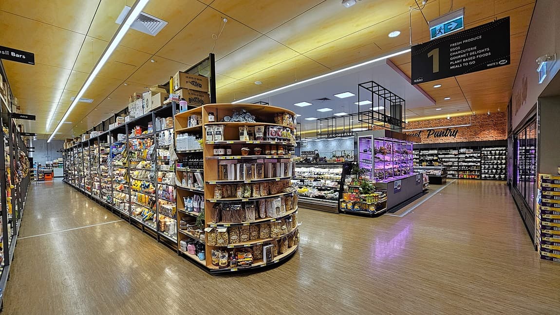 On the left, there are tall shelves stocked with various snacks, cereals, and packaged goods. In the middle, there is an open area with a display of fresh produce and a sign reading "The Pantry" in the background. On the right, there are more shelves with jars and bottles, and a refrigerated section with glass doors. The floor is light-coloured wood, and the ceiling has wooden panels with recessed lighting and ventilation units.