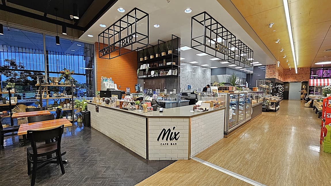 On the left, there are wooden tables and chairs near large windows with metal bars. In the middle, there is a café bar counter with a "Mix Café Bar" sign, displaying various pastries and drinks. The background features shelves with coffee supplies and a brick wall. On the right, there are glass display cases with more pastries and a section with packaged goods. The floor is a mix of dark herringbone tile and light wood, and the ceiling has recessed lighting and black metal frames.