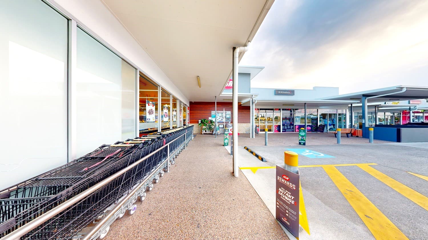 On the left, there is a row of shopping trolleys lined up against a glass storefront. In the middle, there is a walkway with a textured concrete floor and a metal railing. On the right, there is a car park with marked spaces, including yellow pedestrian lines and a few parked cars. The background includes additional store buildings and a covered walkway. The ceiling is flat and white, extending over the walkway area.