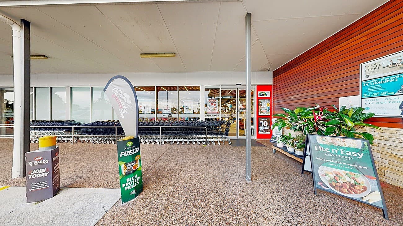 On the left, there is a row of shopping trolleys behind a glass storefront. In the middle, there is a textured concrete floor with a promotional sign for a rewards programme and a flag advertising a product. On the right, there is a wooden wall with posters and a display of potted plants next to a large sign for a meal service. The ceiling is flat and white, extending over the entire area.