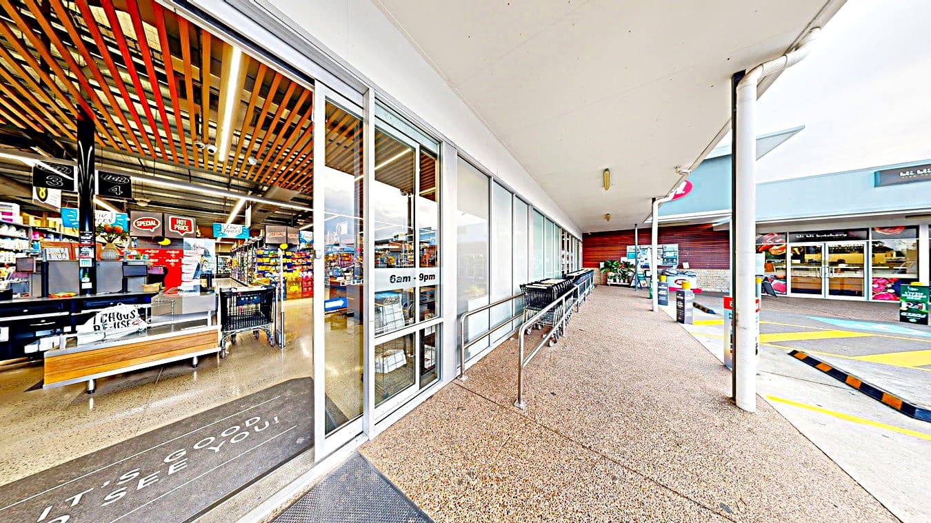 On the left, there is a glass storefront with a sign indicating "6am - 9pm" and a view of the interior with shelves of products. In the middle, there is a walkway with a textured concrete floor and a metal railing. On the right, there is a car park with marked spaces, including yellow pedestrian lines and a few parked cars. The background includes additional store buildings and a covered walkway. The ceiling is flat and white, extending over the walkway area.
