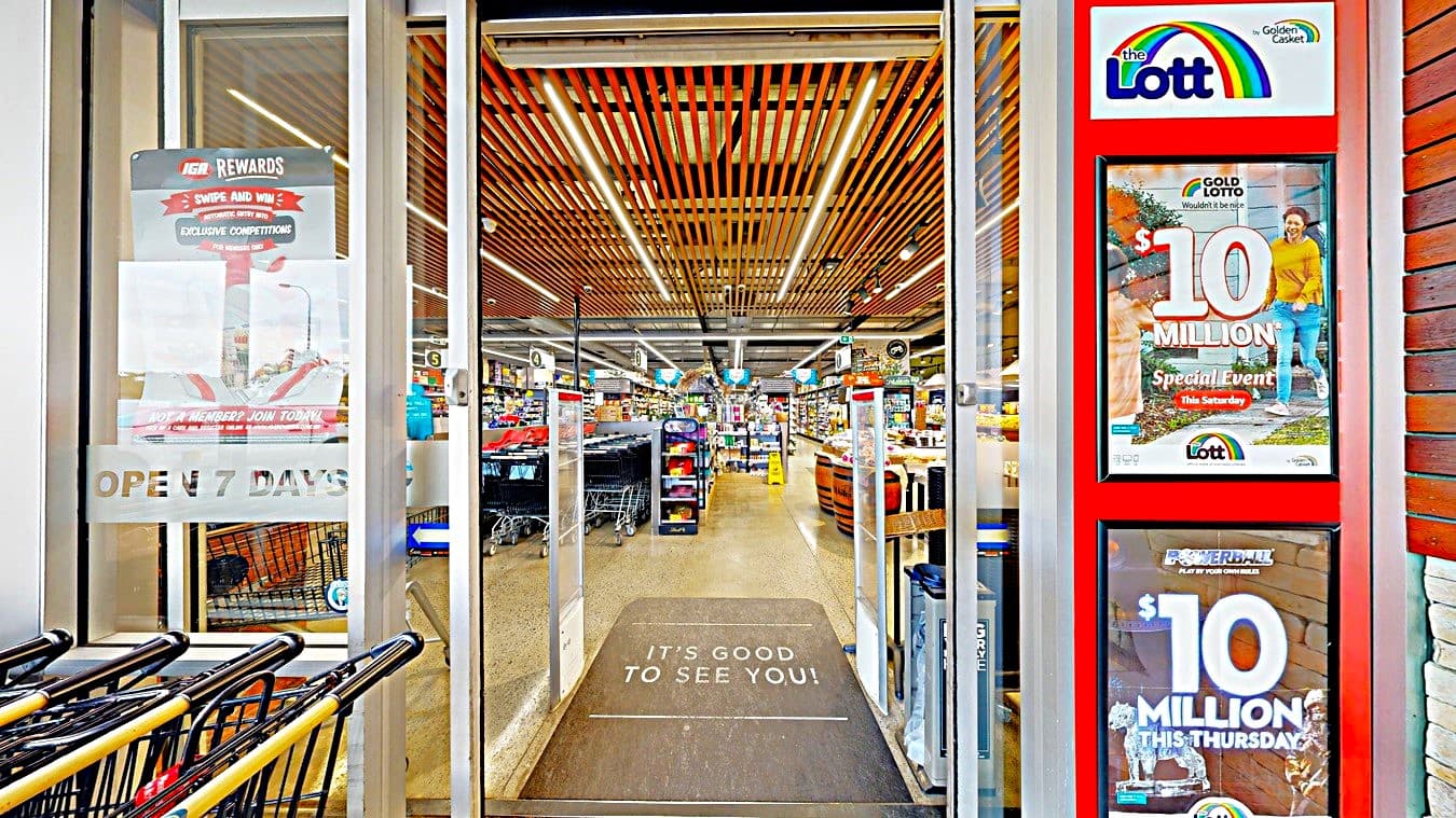On the left, there is a glass door with a sign reading "Open 7 Days" and a row of shopping trolleys. In the middle, there is an entrance mat with the words "It's Good to See You!" leading into a supermarket with aisles of products. On the right, there are two digital screens displaying lottery advertisements. The floor is a mix of concrete and mat, and the ceiling has wooden slats with integrated lighting.