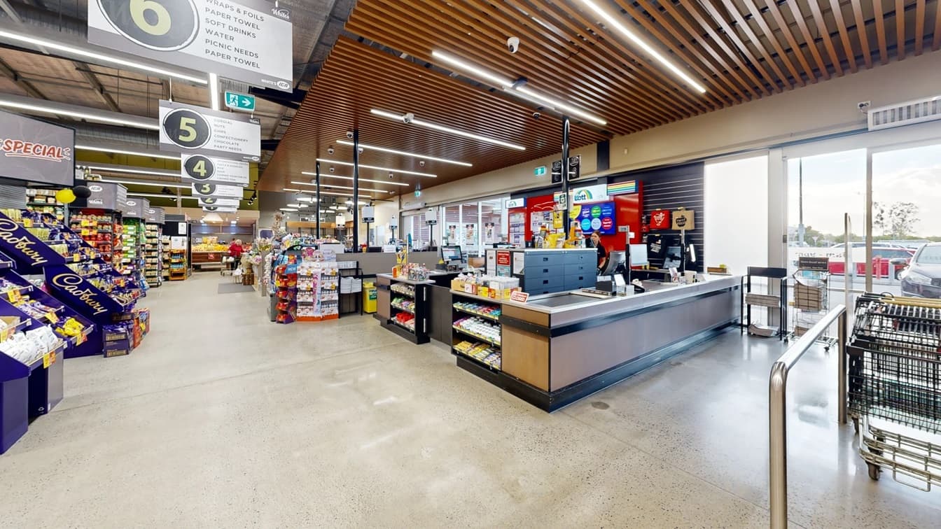 Checkout counter close to aisle no. 6. This is the counter close to the motion-censored glass doors with white-painted metal frames. Noticeable in this image are the shelves for chocolate bars, mints, lollipops, batteries, and lighters. The ceiling has a long, narrow, artificial light and several CCTV cameras that cover different angles of the entire checkout counter area. You can see the cars parked near the store through the glass windows and doors.