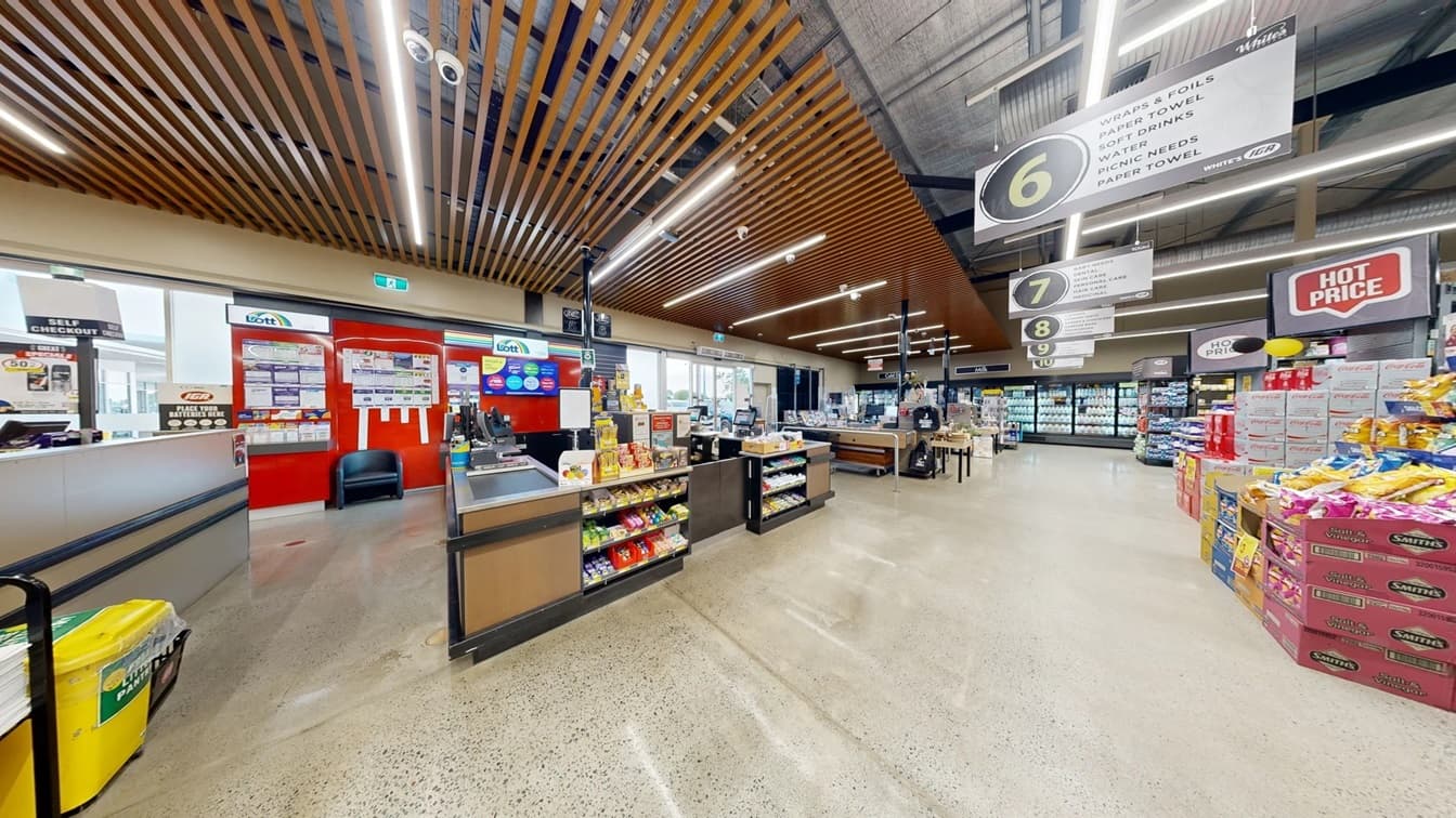 Side view of the grey, wooden with metal strips countertop desk of the customer service section at White’s IGA Bli Bli. Straight ahead from this section is the series of the fridge for milk and cold drinks that occupies most of the internal wall. The ceiling that covers this section has reddish-brown metal strips placed parallel to each other. From this position, square sign boards can be seen hanging on the ceiling above each aisle that are numbered 6-10.