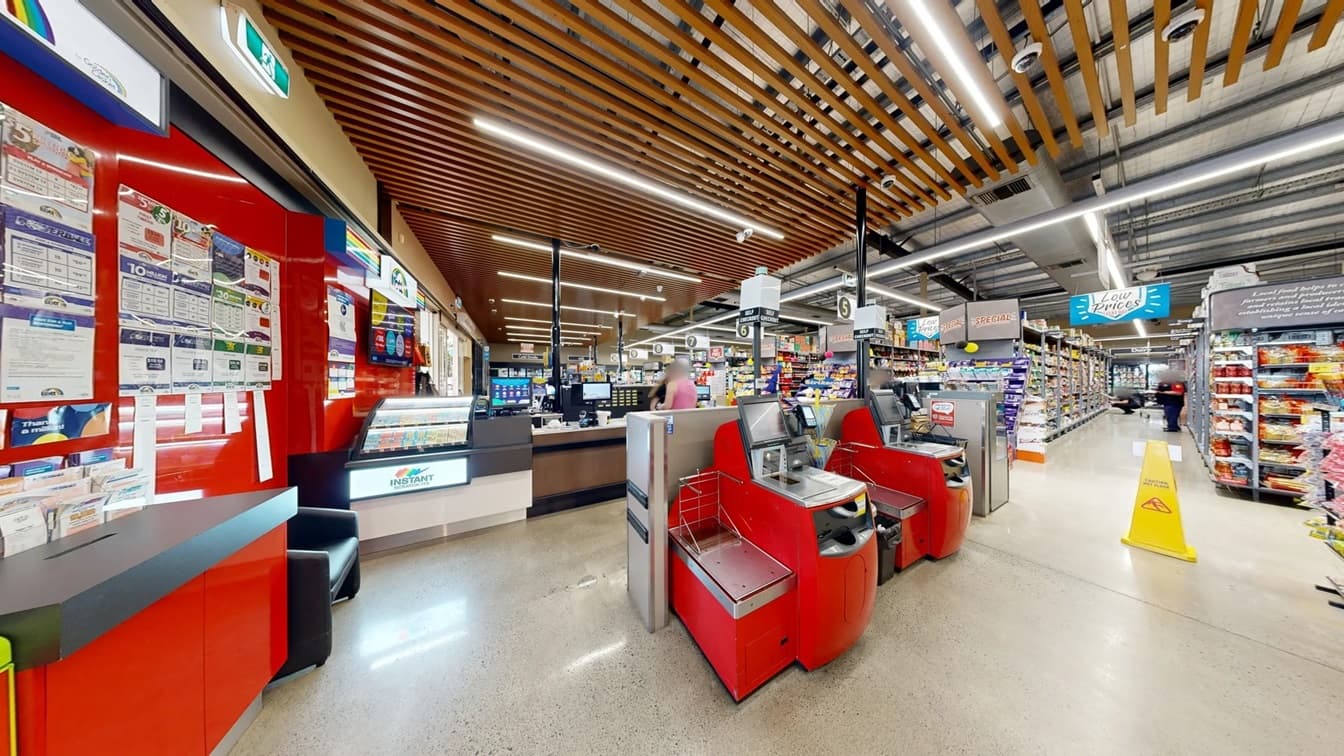 Self-checkout counter at White’s IGA Bli Bli can be identified by the machine’s red color and its small white signboard with light. Batteries, chocolates, candies, and artificial plants are sold on the counters. Placed against the glass walls are black sofa chairs and visible through these walls are the shopping trolleys arranged outside. The accessible car park outside can also be seen from this position.