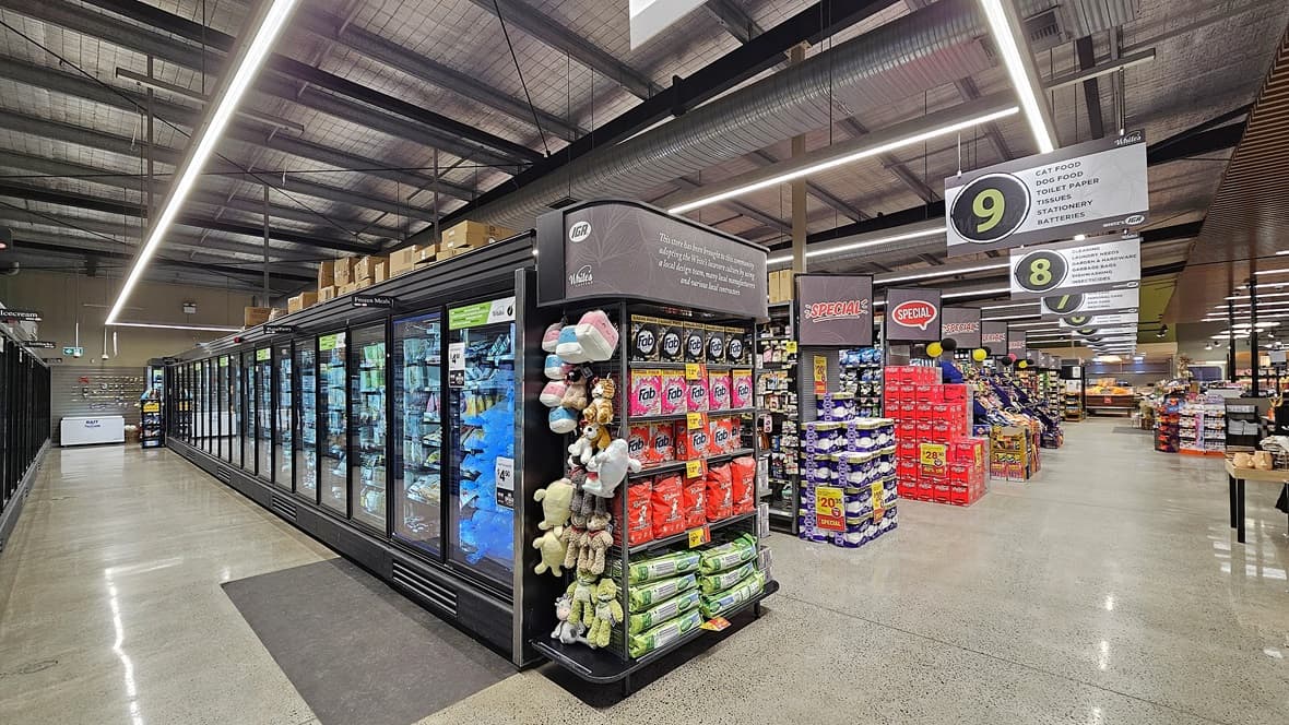 On the left, there are tall refrigerated display cases with glass doors, filled with frozen goods. In the middle, there is a display rack with stuffed animals, snacks, and other small items. In the back, there are more aisles with shelves stocked with various products and a sign indicating checkout lane 9. On the right, there are stacks of packaged goods and additional aisles. The floor is polished concrete, and the ceiling has exposed metal beams and ductwork with linear fluorescent lighting.