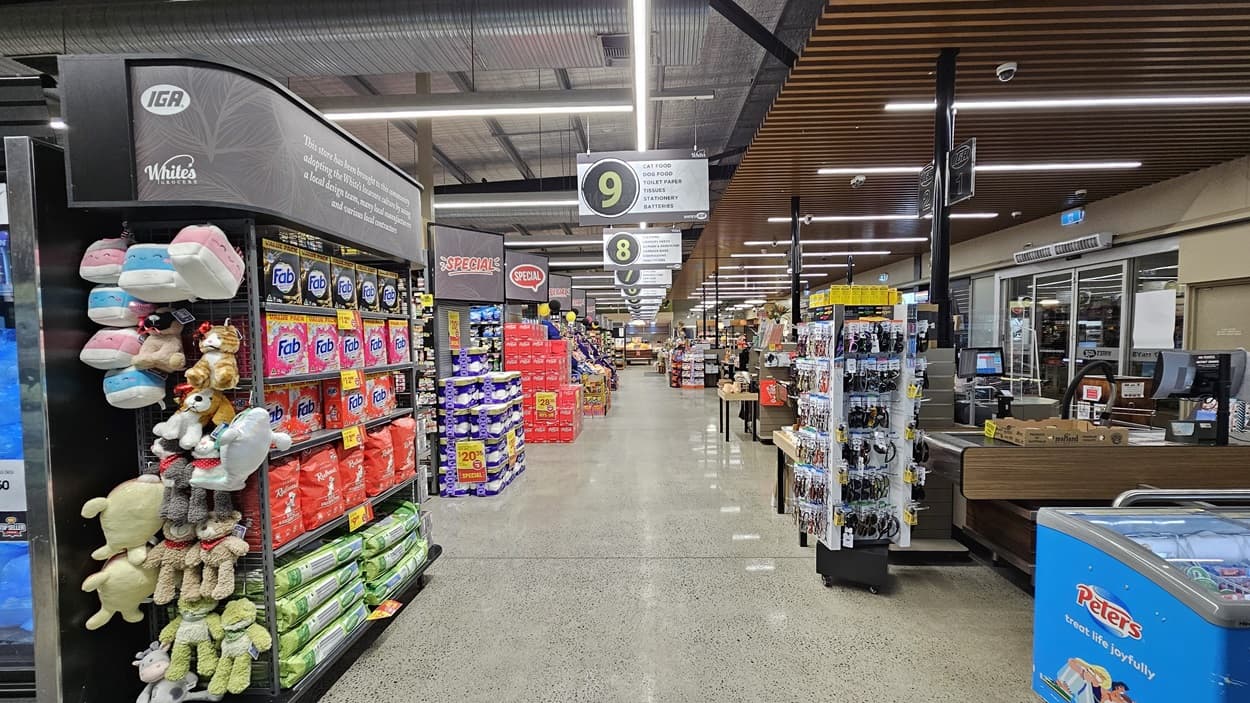 On the left, there are shelves with stuffed animals, pet food, and cleaning supplies. In the middle, there is a wide aisle leading to the background, where checkout lane signs 8 and 9 are visible. The background also has more shelves and displays. On the right, there are checkout counters with a magazine rack and a freezer containing ice cream. The floor is polished concrete, and the ceiling has a mix of wooden panels and exposed metal beams with linear fluorescent lighting.