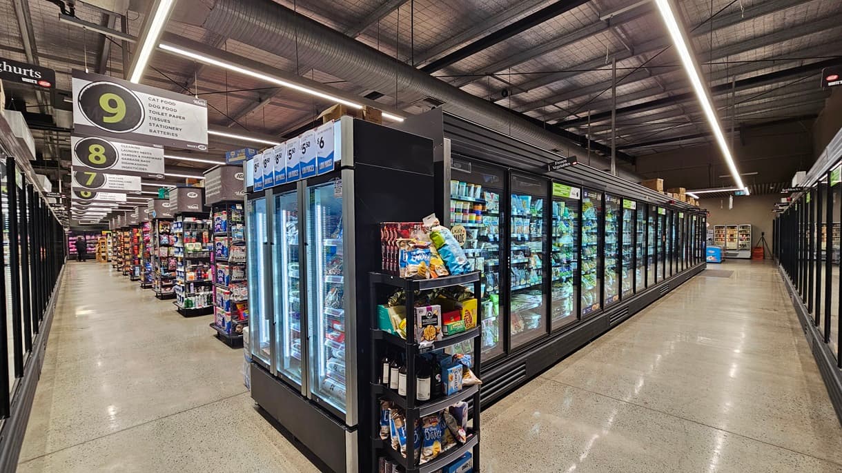 On the left, there are tall refrigerated display cases with glass doors, filled with frozen foods and ice cream. In the middle, there is a smaller refrigerated unit with drinks and snacks, and signs for checkout lanes 7, 8, and 9 hanging above. In the background, more aisles with shelves stocked with various products are visible. On the right, there are additional refrigerated display cases with glass doors, containing frozen goods. The floor is concrete, and the ceiling has exposed metal beams.