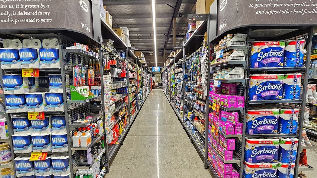 On the left, there are shelves stocked with paper towels, cleaning supplies, and bottled drinks. In the middle, there is a long aisle leading to the background, where more shelves with various products are visible. On the right, there are shelves filled with toilet paper, tissues, and other paper products. The floor is polished concrete, and the ceiling has exposed metal beams with linear fluorescent lighting.