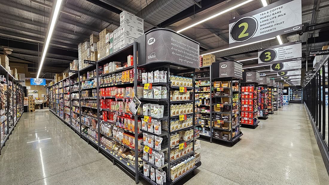 On the left, there are shelves stocked with various packaged goods, including cereals and snacks, with boxes stacked on top. In the middle, there is a wide aisle leading to the background, where more shelves and signs for aisles 2 and 3 are visible. On the right, there are refrigerated display cases with glass doors, containing dairy products and drinks. The floor is polished concrete, and the ceiling has exposed metal beams with linear fluorescent lighting.