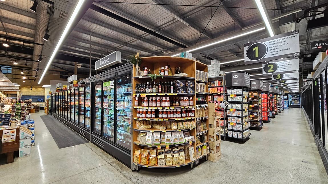 On the left, there are tall refrigerated display cases with glass doors, filled with frozen foods and beverages. In the middle, there is a circular shelf unit stocked with snacks, sauces, and condiments. In the background, there are signs for aisles 1, 2, and 3, with more shelves and products visible. On the right, there are additional shelves with various packaged goods and a sign for sausages. The floor is polished concrete, and the ceiling has exposed metal beams with fluorescent lighting.