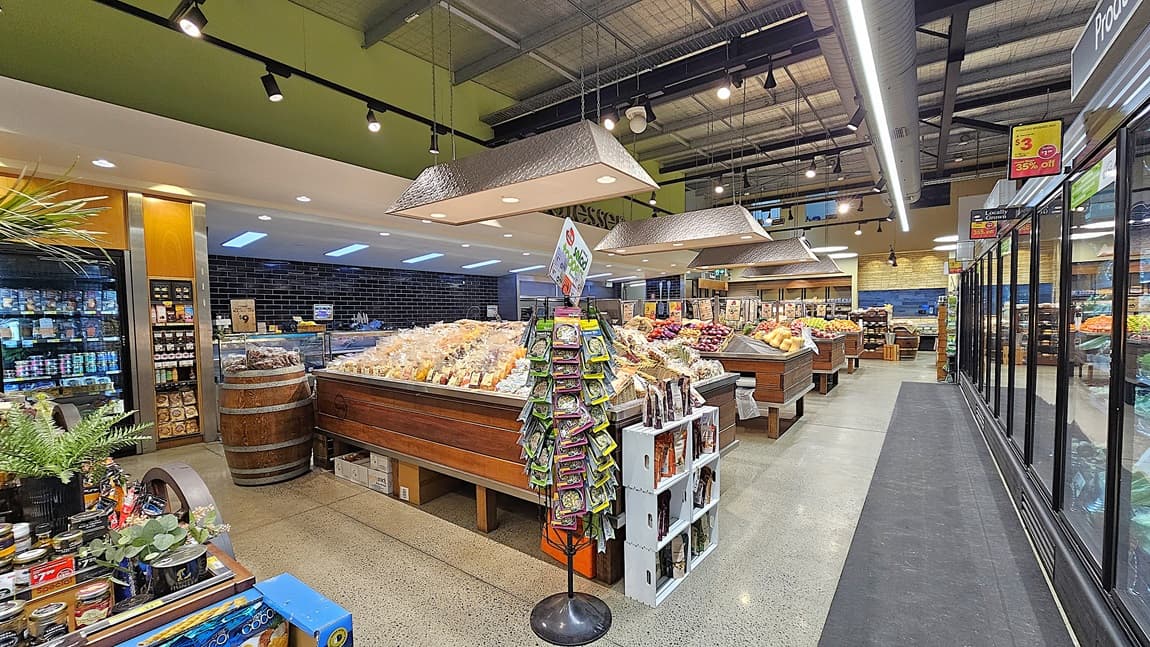 On the left, there are shelves with plants, a wooden barrel, and various packaged goods. In the middle, there is a rotating stand with greeting cards and a white shelf with snacks. In the background, there are wooden produce bins filled with fruits and vegetables, and a sign for groceries. On the right, there are tall refrigerated display cases with glass doors, containing frozen foods. The floor is polished concrete, and the ceiling has exposed metal beams with large rectangular light fixtures.