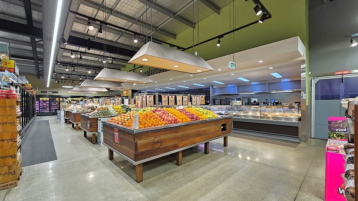 On the left, there are tall refrigerated display cases with glass doors, containing various products. In the middle, there are wooden produce bins filled with oranges, apples, and other fruits, with large hanging light fixtures above. In the background, there are more shelves and a sign for the bakery section. On the right, there is a deli counter with meats and cheeses, and shelves with pet food. The floor is polished concrete, and the ceiling has exposed metal beams with track lighting.