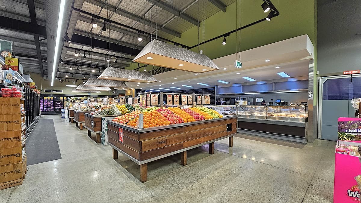 On the left, there are shelves with packaged goods and a refrigerated section with glass doors. In the middle, there are wooden produce bins filled with various fruits, including oranges, apples, and pears, with large rectangular light fixtures hanging above. In the background, there is a bakery section with display cases and a sign. On the right, there is a deli counter with meats and cheeses, and a pink display stand with pet food. The floor is concrete, and the ceiling has exposed metal beams