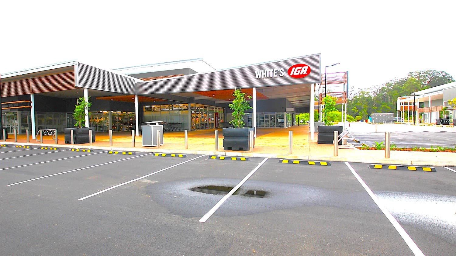 A supermarket entrance with a large "IGA White's" sign in the centre. On the left, there are parking spaces and a covered walkway with wooden slats. On the right, more parking spaces and a glass storefront. In the middle, a pedestrian crossing leads to the entrance. The background features trees and additional buildings. The foreground has a mix of asphalt and concrete surfaces, with marked parking spaces, including accessible spots.