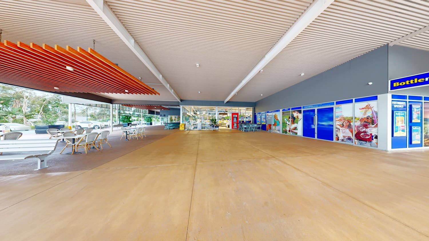 On the left, there are outdoor tables and chairs under a wooden slatted ceiling. On the right, there are blue storefronts with large windows displaying advertisements. In the middle, a wide walkway leads to the supermarket entrance with glass doors. The background includes trees and additional seating. The floor is a smooth, light brown concrete, and the ceiling above the walkway is white with exposed beams.