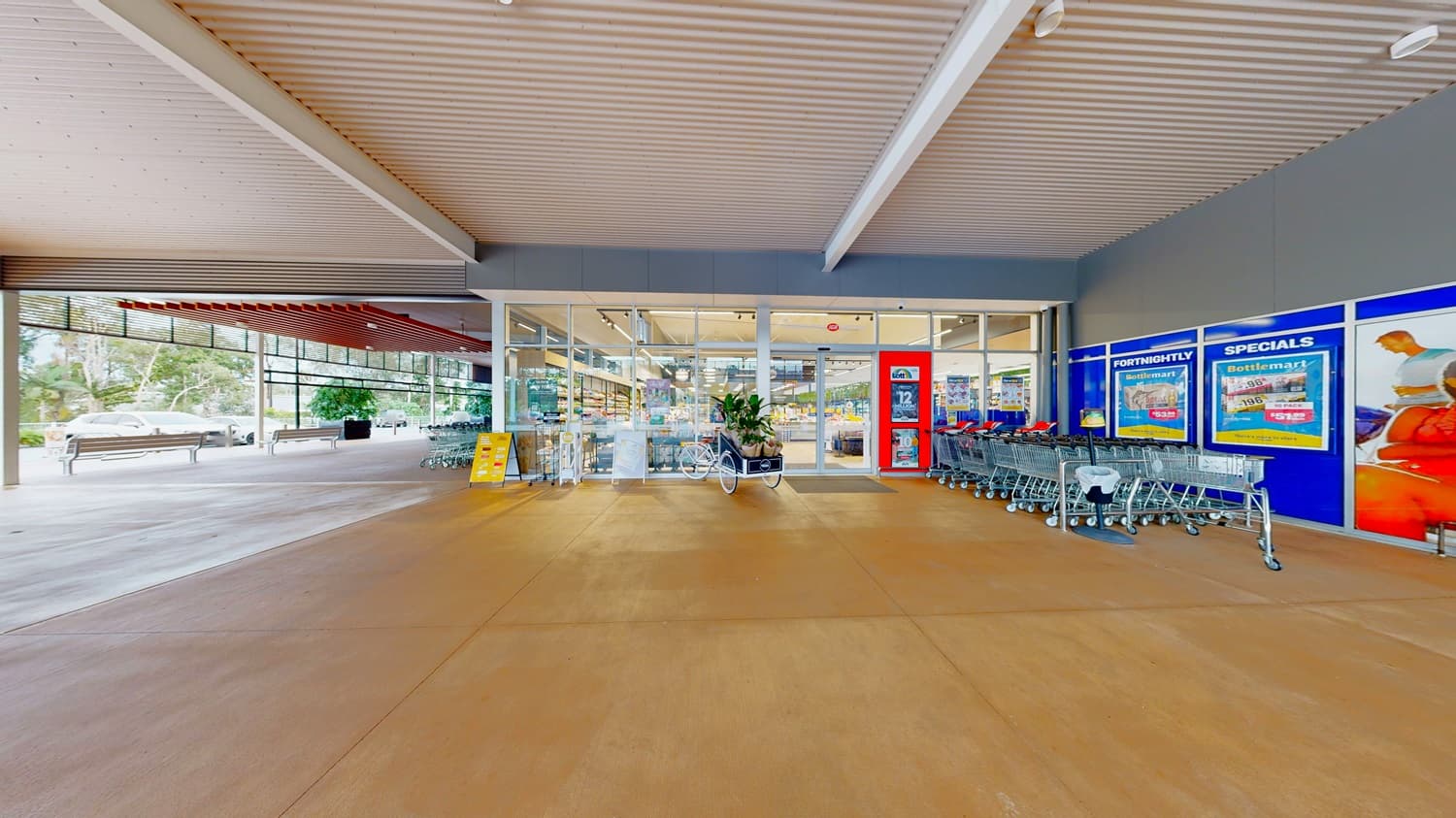 On the left, there is a covered walkway with a wooden slatted ceiling and a parked car in the distance. On the right, there are blue storefront windows displaying advertisements and a row of shopping trolleys. In the middle, glass doors lead into the supermarket entrance. The background includes trees and additional seating. The floor is smooth, light brown concrete, and the ceiling above the walkway is white with exposed beams.