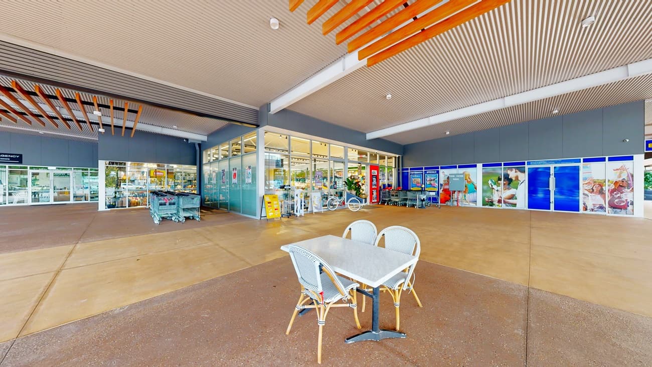 On the left, there are glass doors leading into a store, with shopping trolleys nearby and a wooden slatted ceiling above. On the right, there are large blue storefront windows with various advertisements and a few tables and chairs. In the middle, there is a small table with two chairs. The background includes more glass doors and windows. The floor is a mix of smooth light brown and dark brown concrete, and the ceiling is white with wooden slats.