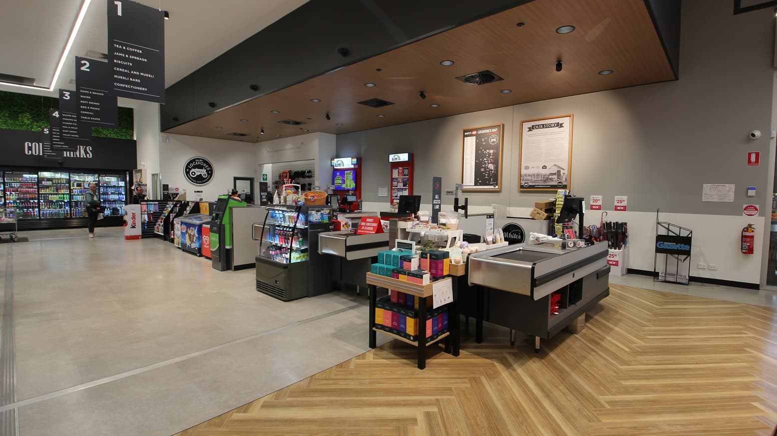 A view of the checkout area which features three cash registers whilst further at the back is the express check out area. A range of products is on display leading up to the check out area including a small fridge of cold drinks, ice cream freezers, and an ATM. Along the back wall is signage as well as marketing materials for ‘Lotto’. Further to the right is a fire extinguisher located on the wall as well as red signage indicating the location of emergency equipment.