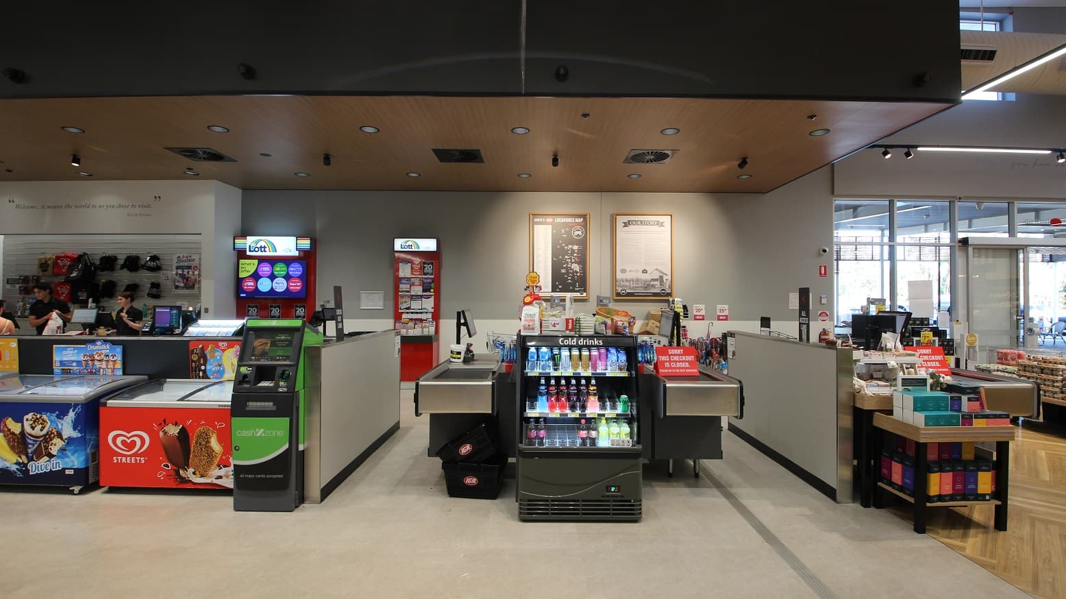 A direct view of three cash register aisles, with two ice cream freezers situated to the left next to an ATM and marketing materials for ‘Lotto’ can be seen towards the back. To the left three people can be seen chatting near the express check out area. To the right is a small display featuring boxes in a range of colours including green, yellow and orange. Directly ahead is a small drinks fridge and on the far back wall are two large prints with brown frames.