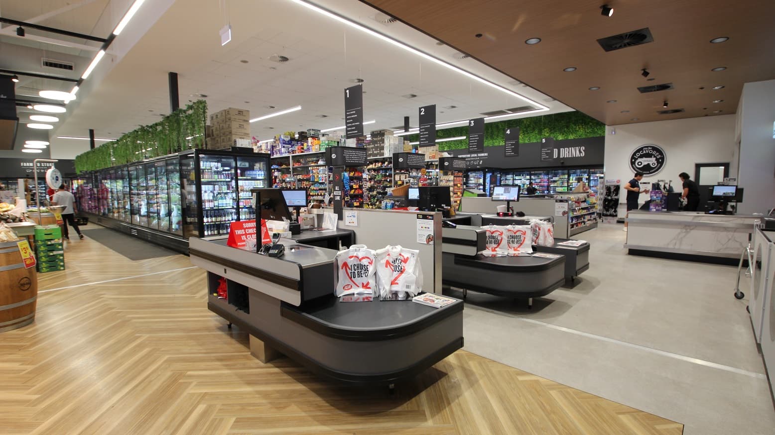 A view from near the entrance back towards the check out area. To the left is a range of fruit and vegetables on display and in the fridge. Directly in front is one of the cash registers with a red sign stating this register is currently closed. Red, white and black plastic bags are hanging from a small stand. Behind are the 5 grocery aisles. To the right is the express check out area where two people dressed in black can be seen talking. On the wall above these people is a 'Locovare' sign.