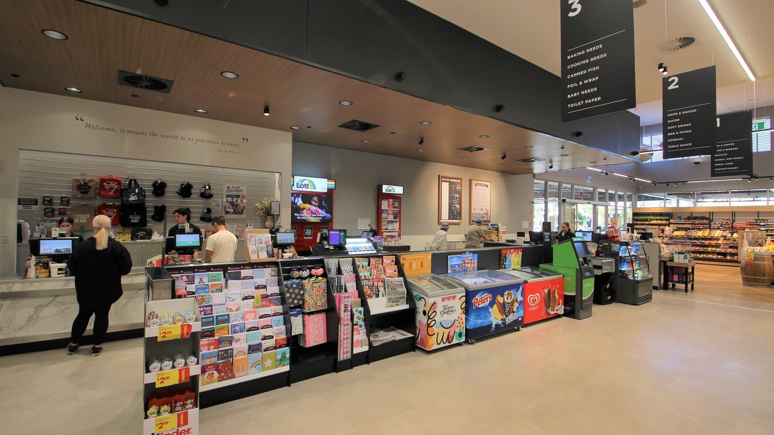 Directly ahead to the left is a lady dressed in black being served by another lady at the express check out area. On the back wall is a range of bags and other merchandise as well as marketing materials for ‘Lotto’ on display to the right. In front of this express checkout area is a range of chocolates, a display of gift cards, wrapping paper and ribbons next to the newspaper and magazine stand. Next to three ice cream freezers and an ATM leading up to the three cash register aisles.