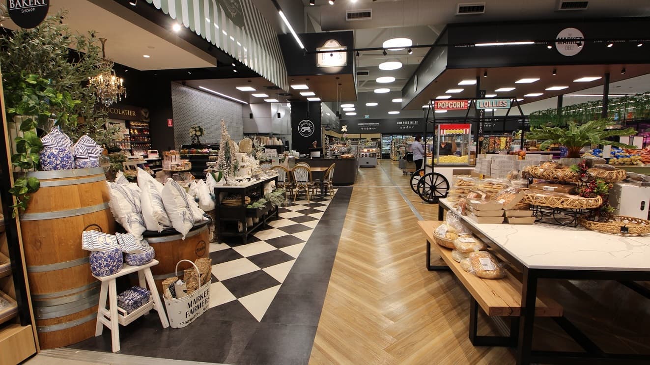 A brightly lit space with a range of homewares on display to the left including white cushions, home decor and floral arrangements. To the right, separated by a wide walkway is a table and bench with a range of snacks in boxes and trays on display. A popcorn stand is further along to the right situated next to a ‘Lollies’ stand. The walls to the left have black and white patterned tiles with shelves and is located next to a range of chocolates with a sign stating ‘Chocolatier’.