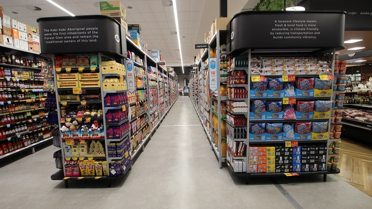A view down one of the aisles featuring a range of grocery items. To the left of the entry to this aisle is a display featuring a range of chocolates including ‘Toblerones’. To the right of the entry to this aisle is a display of soft drinks, biscuits and ‘Zooper Doopers’.