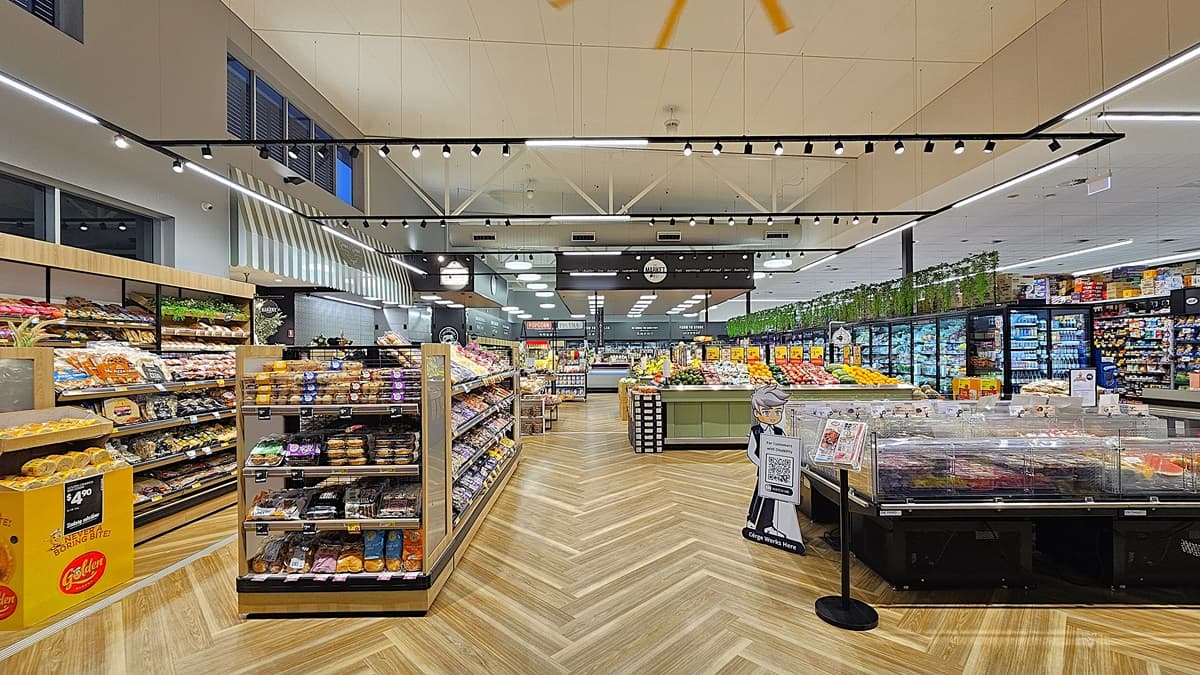 A supermarket interior with a wooden chevron-patterned floor. On the left, there are shelves filled with baked goods and snacks. In the middle, there is a large island with fresh produce and a cardboard cutout of a person holding a sign. The ceiling is high with exposed beams and track lighting. On the right, there are refrigerated sections with glass doors containing meats and dairy products. The background features more shelves and a section with potted plants on top.