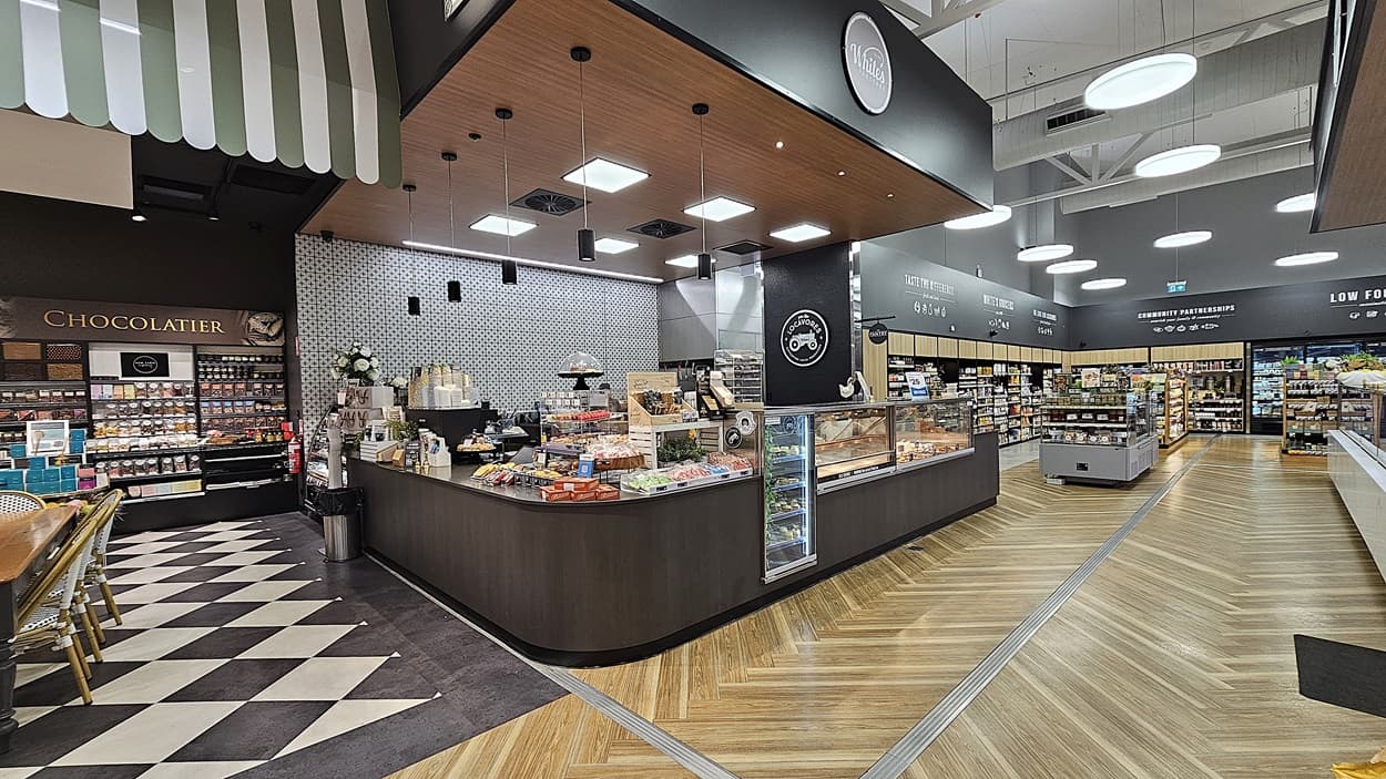 A supermarket with a mix of black-and-white chequered and wooden chevron-patterned floors. On the left, there is a chocolaterie counter with various chocolates and pastries. In the middle, there is a large island with a bakery section, including cakes and bread. The ceiling has wooden panels with square lights and exposed white beams. On the right, there are aisles with shelves stocked with groceries and a refrigerated section in the background.