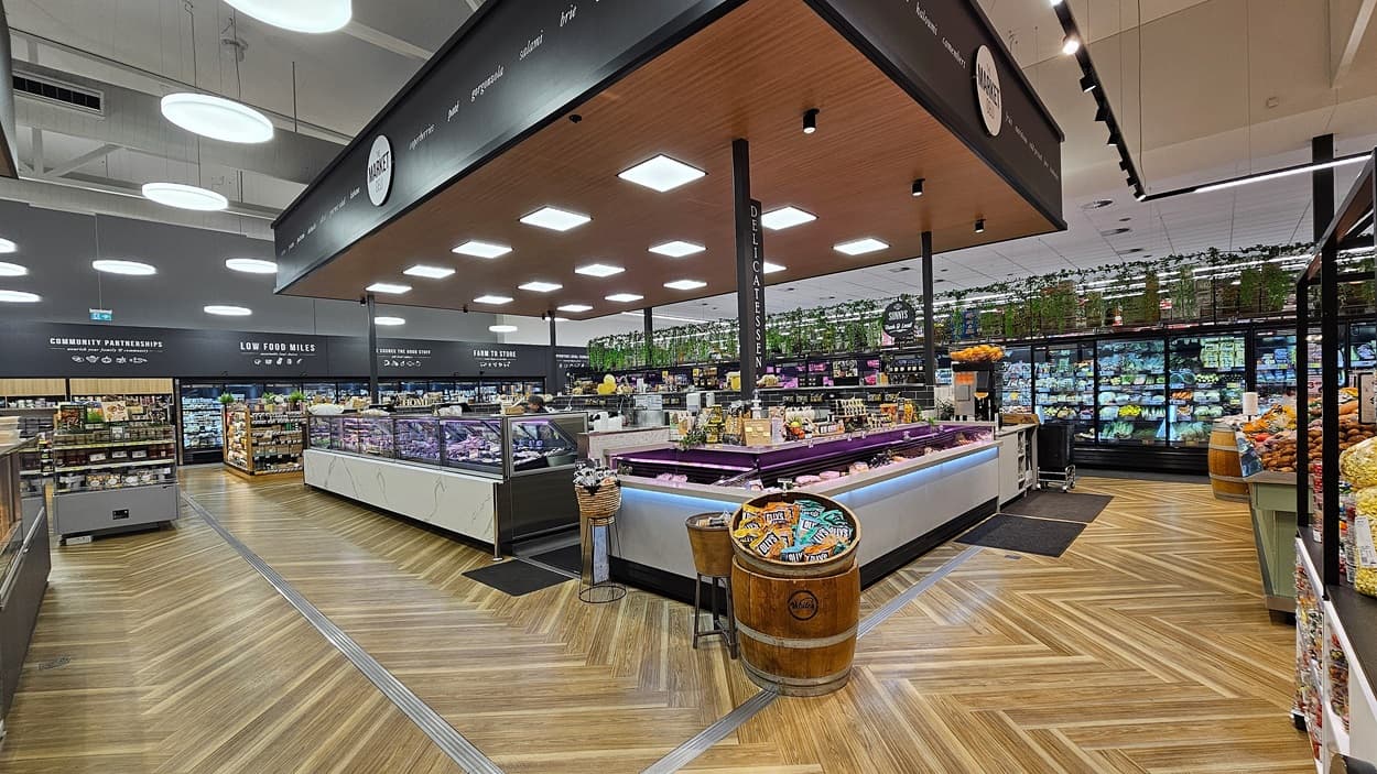 A supermarket with a wooden chevron-patterned floor. On the left, there are shelves with various packaged goods and a sign for gourmet ingredients. In the middle, there is a large island with refrigerated display cases containing meats and cheeses, topped with a wooden ceiling and square lights. On the right, there are refrigerated sections with glass doors displaying dairy products and vegetables. The background features more shelves and a section with potted plants on top.