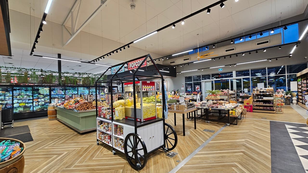 A supermarket with a wooden chevron-patterned floor. On the left, there are refrigerated sections with glass doors displaying fresh produce and a green display table with fruits and vegetables. In the middle, there is a black popcorn cart with a red sign and a variety of snacks. The ceiling is high with white panels and black track lighting. On the right, there are more display tables with baked goods and packaged items, and a section with potted plants on top of shelves.