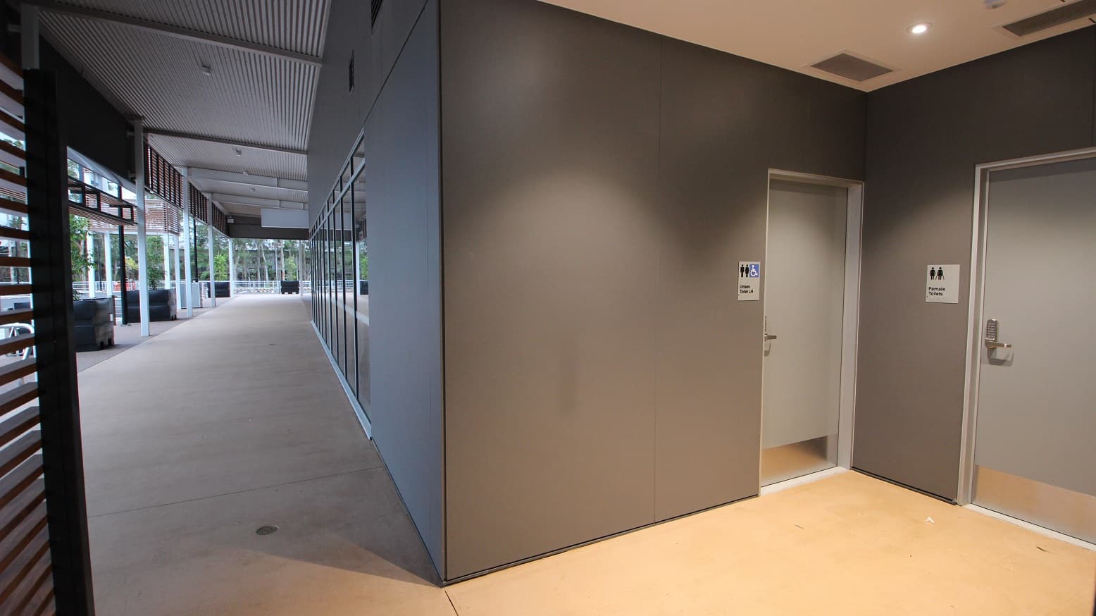 To the left of the picture is a view of the car park featuring the pot plants. To the right are grey walls and a beige floor leading into the female toilets and the Unisex accessible toilet. Both of these doors are light grey in colour and feature silver door handles with a pass code entry.