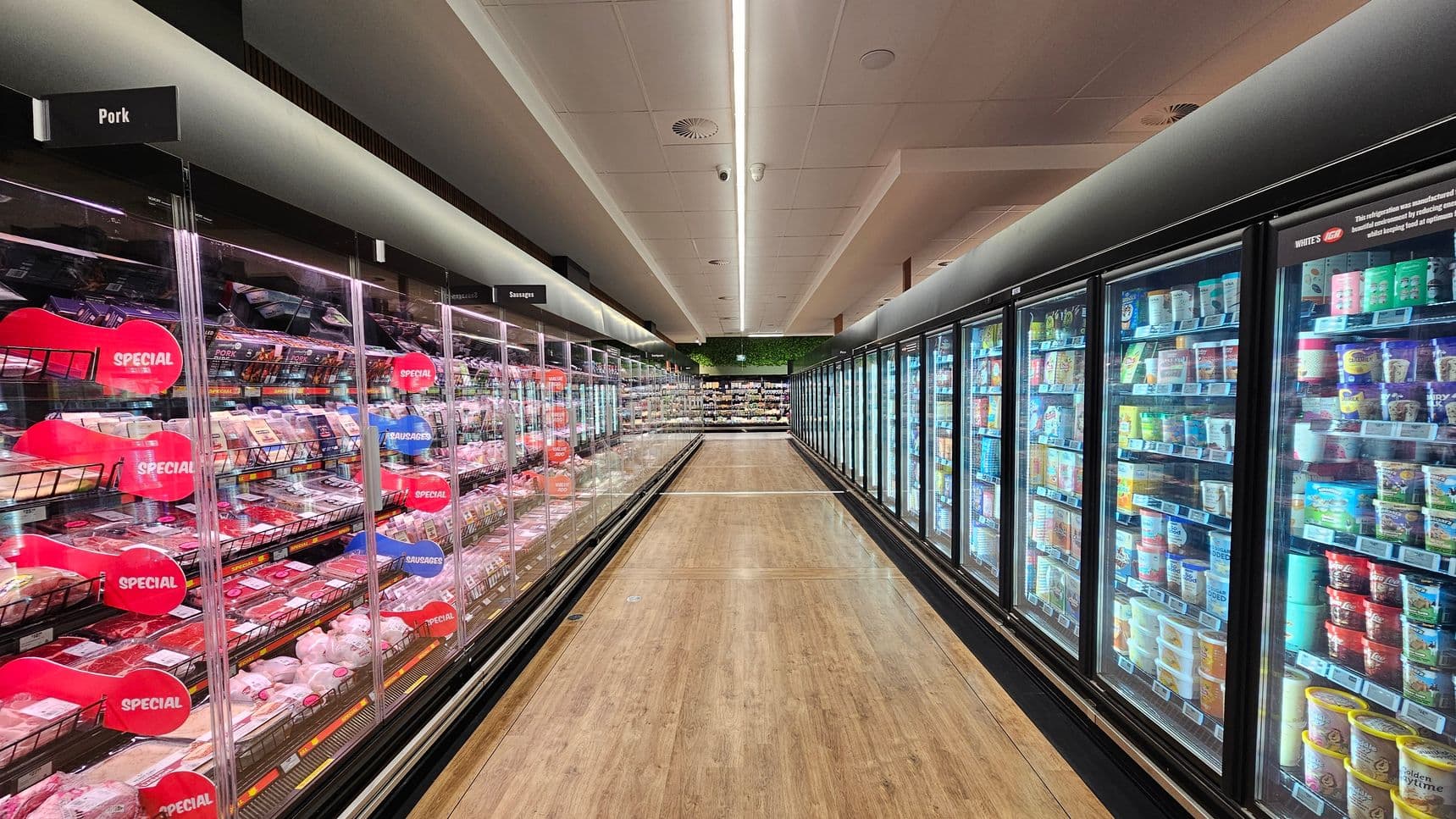 A supermarket aisle with refrigerated sections on both sides. On the left, shelves display various meats, labelled "Pork" and "Sausages," with red "Special" signs. On the right, shelves are stocked with assorted dairy products and ice creams. The floor is wooden, and the ceiling features recessed lighting. At the end of the aisle, more products are visible, with a green plant wall above.
