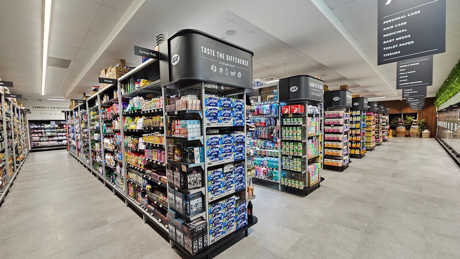 A supermarket aisle with shelves on both sides filled with various products. On the left, shelves are stocked with cleaning supplies and garbage bags. In the centre, a display features paper products like tissues. On the right, more shelves hold personal care items. Overhead signs indicate different sections, and the floor is light-coloured. The ceiling has fluorescent lighting, and the background shows more aisles and a section with plants.