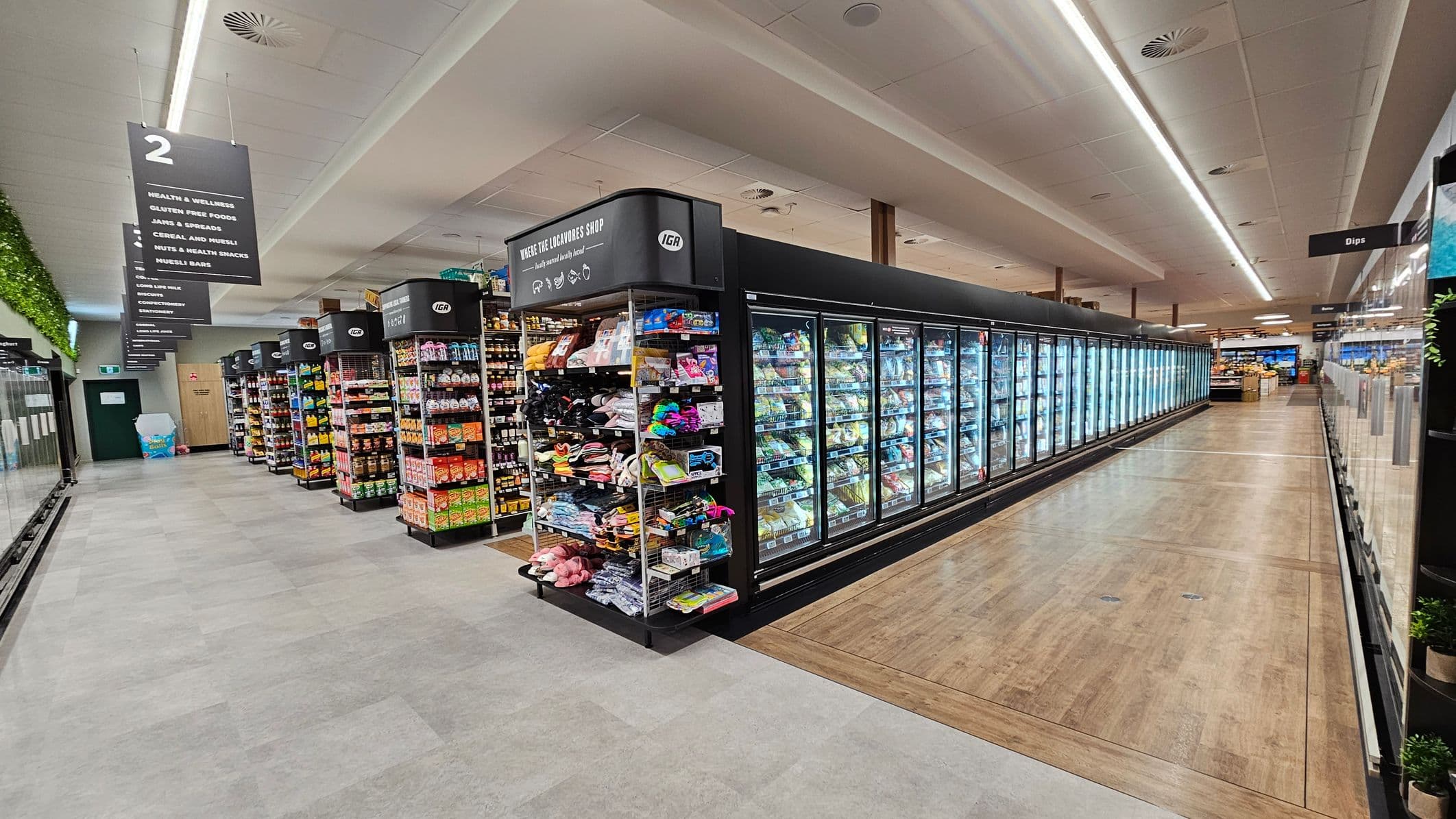 A supermarket aisle with a grey tiled floor and a ceiling with recessed lighting. On the left, there are shelves stocked with various packaged goods and a sign indicating aisle 2, listing categories like health and wellness. On the right, a long row of refrigerated glass doors displays frozen items. The middle of the aisle has a wooden floor section. In the background, more aisles and products are visible, with signs hanging from the ceiling.
